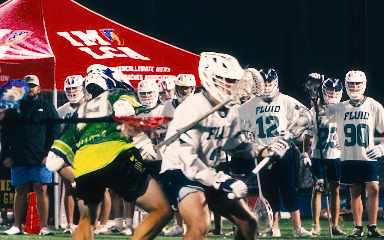 Fluid Lacrosse players in a game at the IMLCA Coaches Summit, with the team watching from the sideline